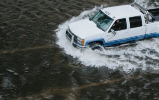 White Chevy Pickup Driving Through A Flooded Road. The Driver peers at the camera warily