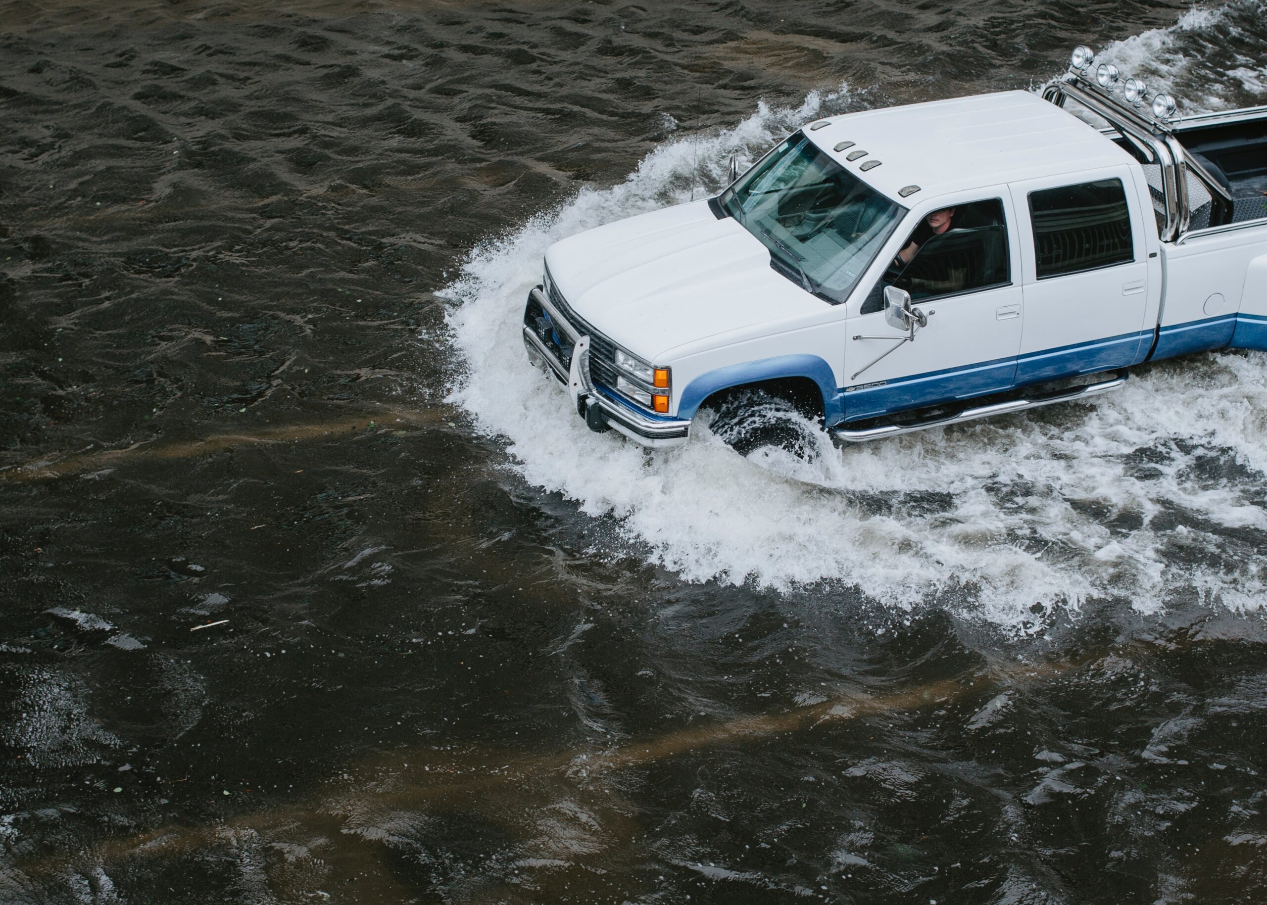 White Chevy Pickup Driving Through A Flooded Road. The Driver peers at the camera warily