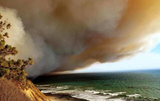 Thick brown smoke billows from a wildfire off of a cliff from the left with a contrasting blue ocean to the right.