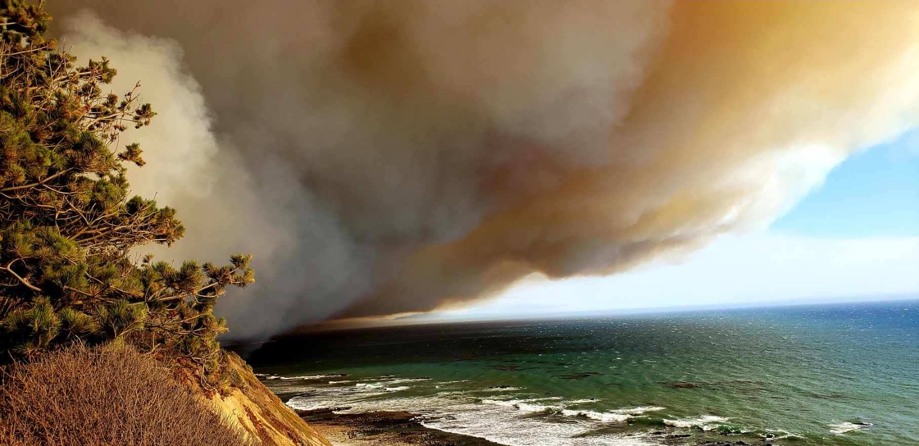 Thick brown smoke billows from a wildfire off of a cliff from the left with a contrasting blue ocean to the right.