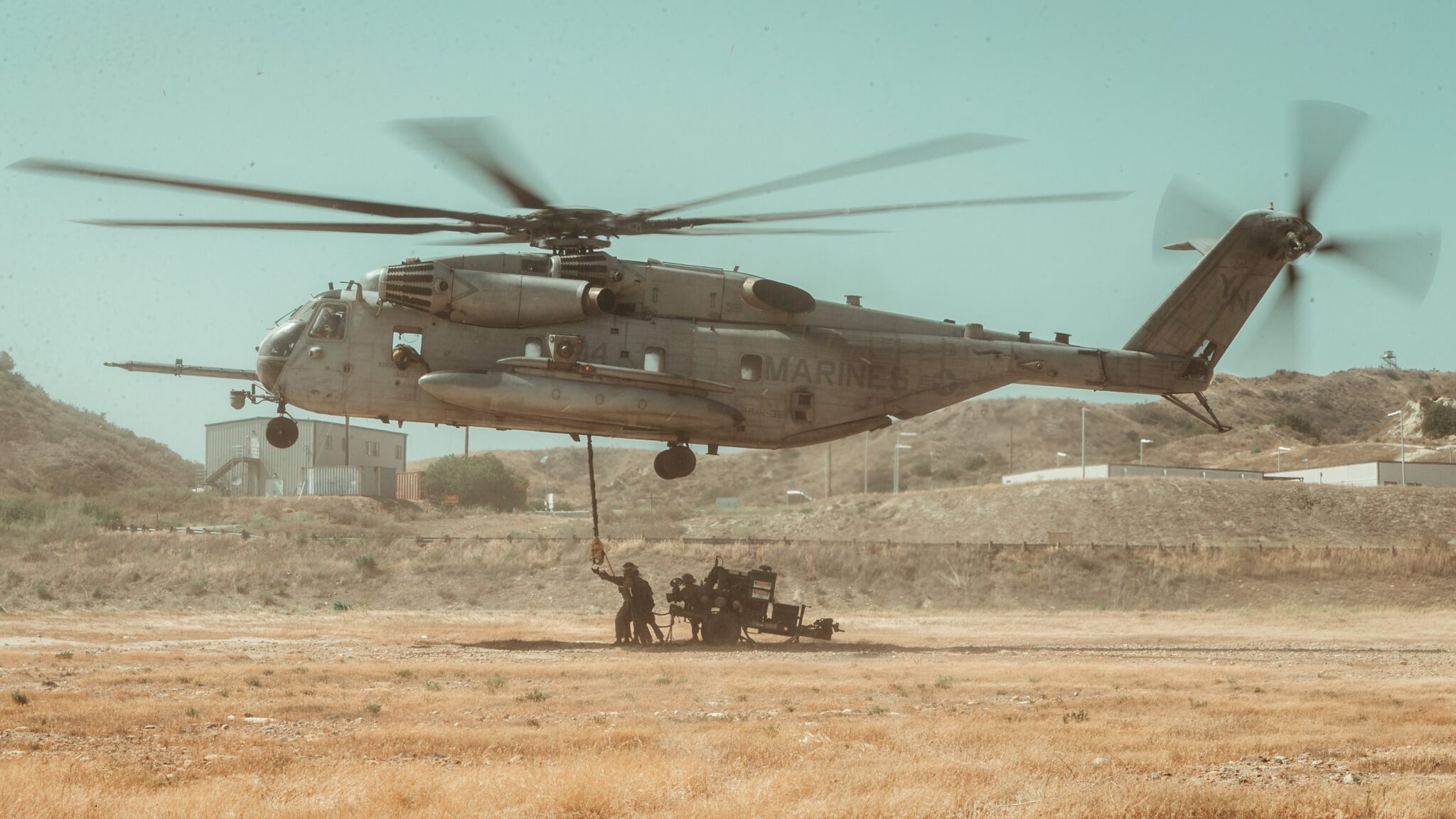 Marines loading ordinance under a hovering helicopter in an open field at a Marine Base