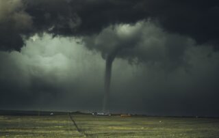 Dark and moody picture of a massive stormy sky over a small Wyoming homestead with a wide field in the foreground. All connected my a tornado spout