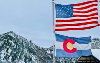 Colorado State Flag waving beneath a US frag with colorado snow-capped mountains in the background.