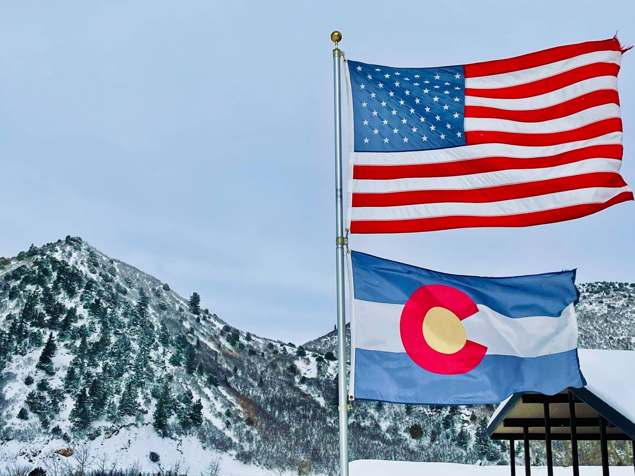 Colorado State Flag waving beneath a US frag with colorado snow-capped mountains in the background.