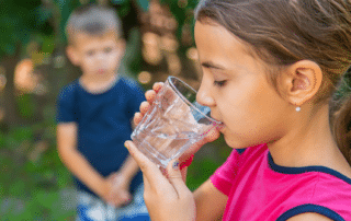 A child drinks water from a clear plastic cup with another child in the background
