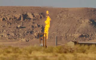 A lone column of burning methane ascends from a dry grassy area with a rocky hill in the background