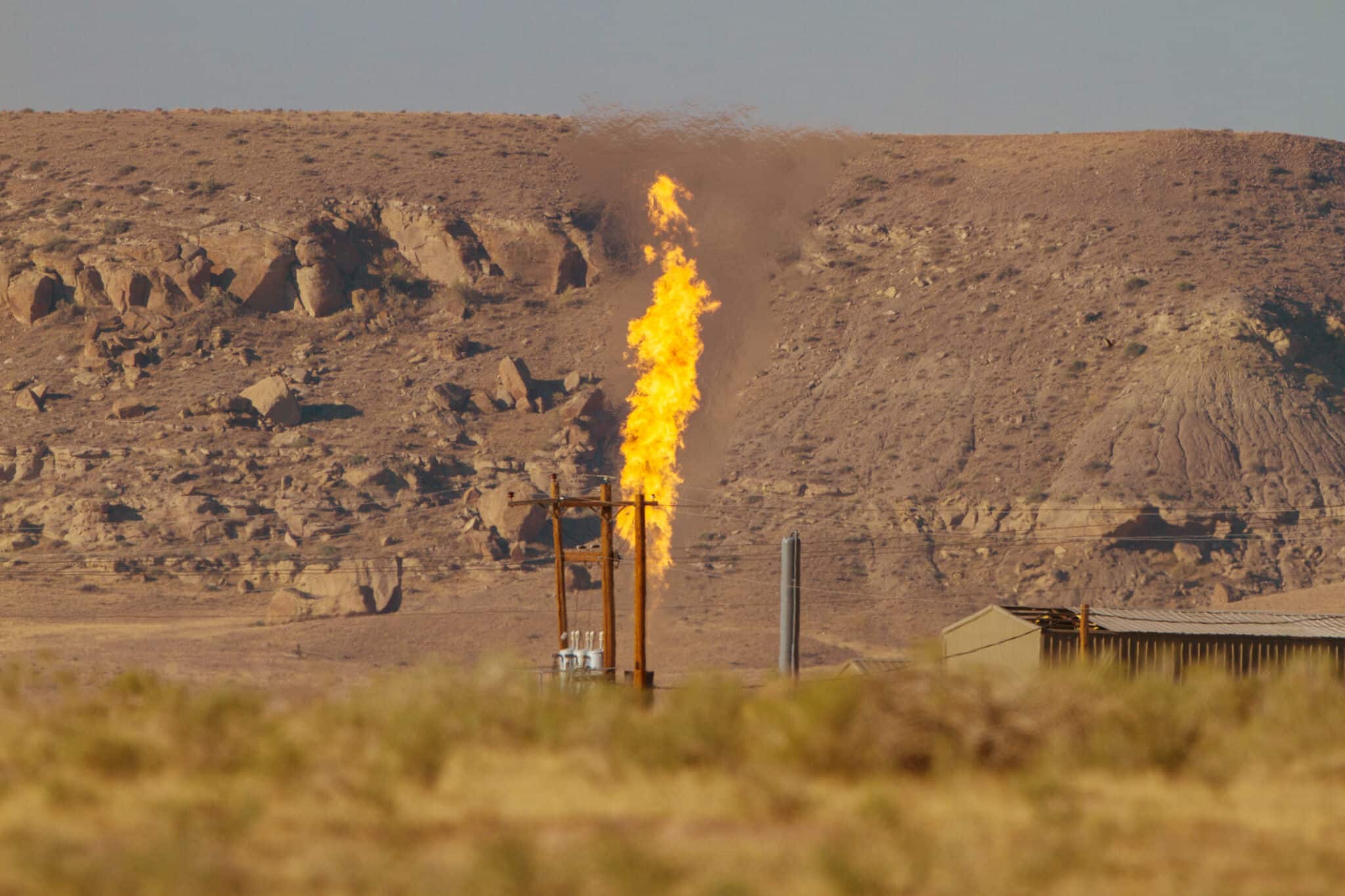 A lone column of burning methane ascends from a dry grassy area with a rocky hill in the background