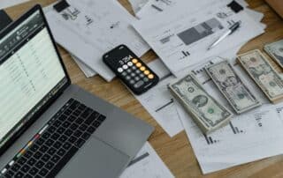 Image of stacks of cash, with stacks of paper, a calculator and a laptop
