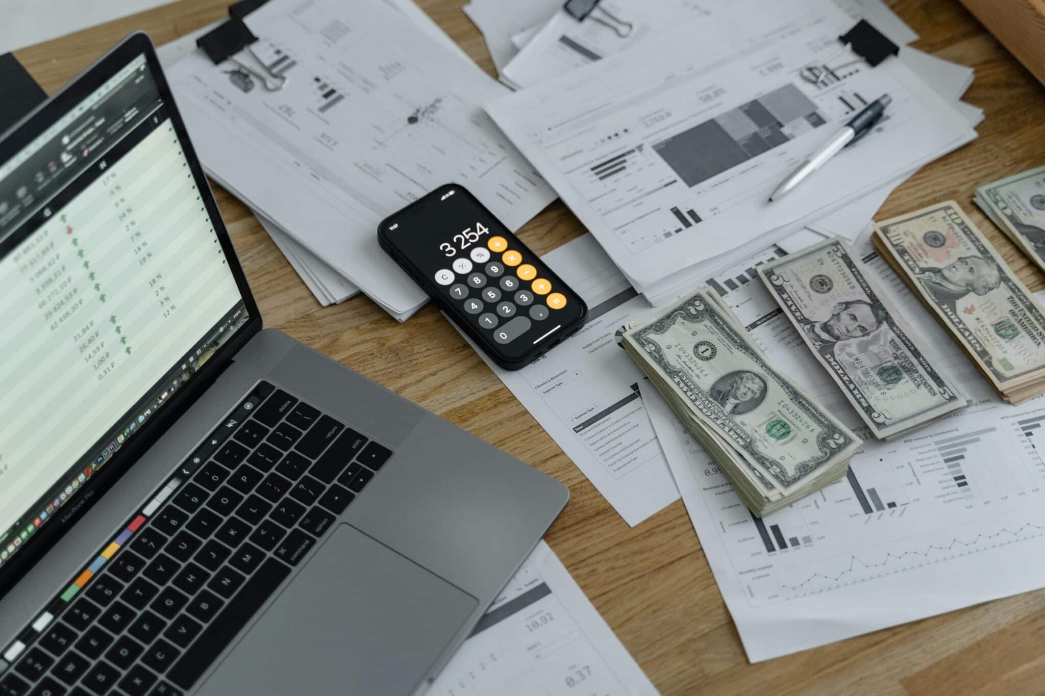 Image of stacks of cash, with stacks of paper, a calculator and a laptop