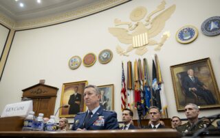Military officials face an unseen committee with the US seal and US Armed Forces colors in the background.