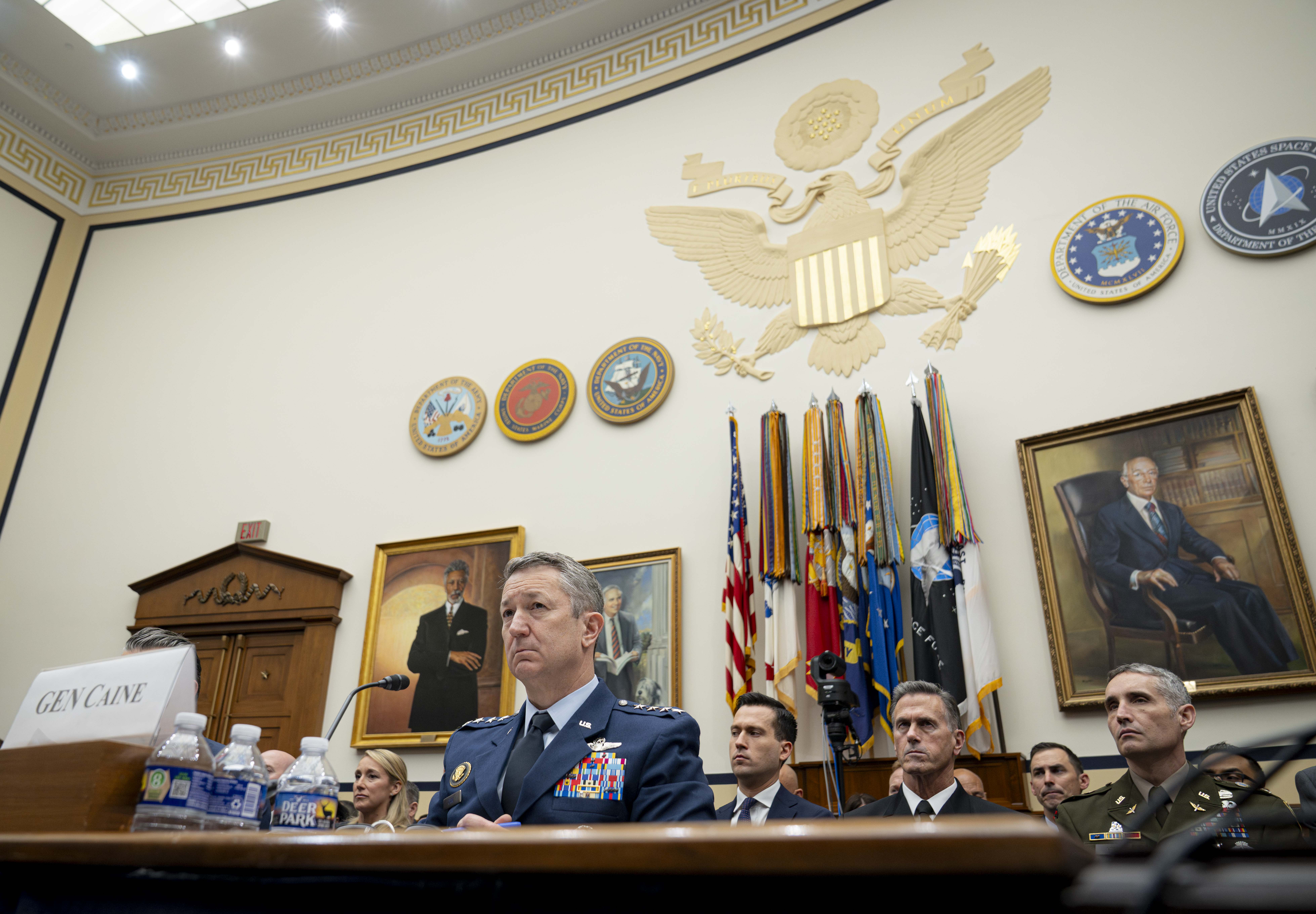 Military officials face an unseen committee with the US seal and US Armed Forces colors in the background.